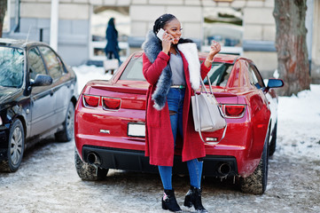 Rich african american girl in red coat and fur against red muscle car speaking on mobile phone....
