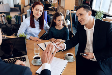 Red-haired mother with daughter watches as cheerful father hands over money to man sitting at table.