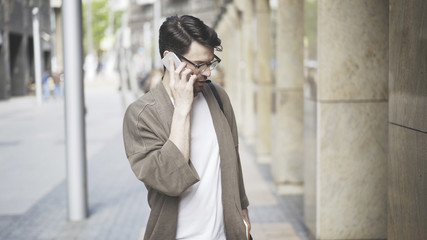 A young serious man dressed in casual clothes talking on the phone on the street. A young man wearing black glasses