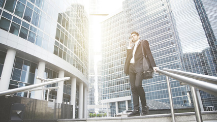 A young businessman dressed in a black suit is talking on the phone on the stairs. Downtown and city on the background