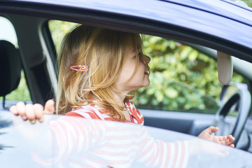  Little girl playing inside car and sitting on front driver seat during a break on a family vacation road trip. Traveling by car with kids. Girl looking at herself in the mirror in the sun blind