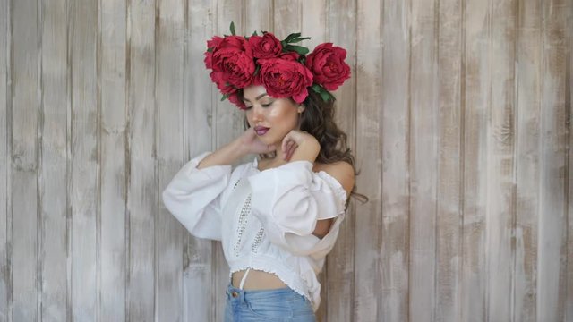 Girl posing in front of camera. young woman in a wreath of scarlet peonies on her head, dark long curly hair descends on the shallow shoulders.