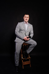 Handsome man in gray suit with microphone against black background on studio sitting on chair. Toastmaster and showman.