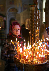 Praying young woman with candle near pedestal with many other candles in church by her hand to show her faith and esteem to God