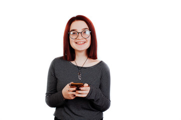 Cheerful young woman with short red hair wearing glasses typing message on phone and looking at...