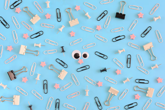 White paper clips, clerical buttons and pink stars are randomly scattered on a blue background. Funny smiley from clips in the center. A picture photographed from above.