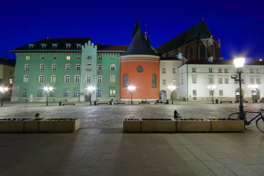 Mały Rynek w Krakowie, Polska, nocny widok, zadbane zabytkowe kamienice, dach bazyliki mariackiej w tle, świecą uliczne lampy, ludzie relaksują się na ławkach, piękne ciemnoniebieskie niebo