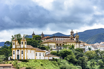 Ancient historical churches among the houses and streets of Ouro Preto city in Minas Gerais with hills and clouds in the background