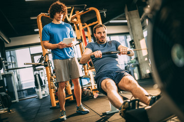 Young man with his personal instructor in fitness center. Using rowing machine for cardio training...