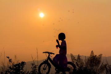 Fototapeta na wymiar Silhouette little girl sitting on bicycle playing with bubble wand on mountain at sunset background