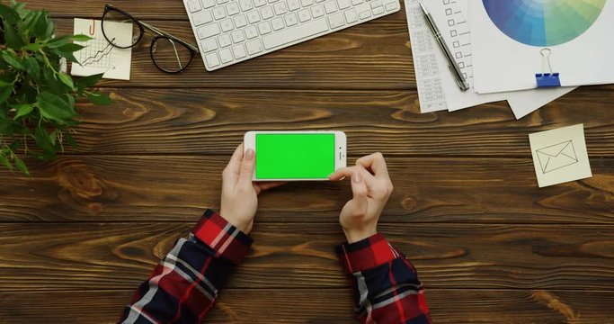 View above on the female hands with a red plaid sleeves working on a white smartphone with a green screen horizontally on the wooden table with a keyboard, charts and office stuff. Chroma key.