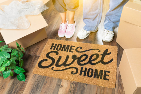 Man and Woman Standing Near Home Sweet Home Welcome Mat, Moving Boxes and Plant