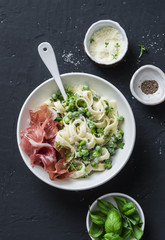 Fettuccine pasta with creamy sauce, green peas and prosciutto on dark background, top view