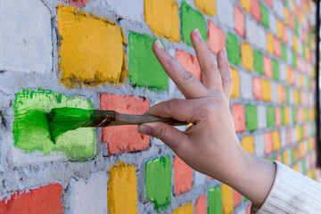 Obraz premium Young Caucasian shapely woman paints a brick wall with paint brush in different colors. Red, blue, yellow, green. Capacity with paint and hands close-up view