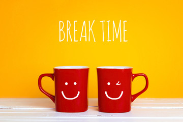 Two red coffee mugs with a smiling faces on a yellow background with the phrase Break time.