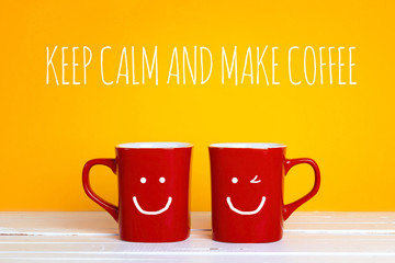 Two red coffee mugs with a smiling faces on a yellow background with the phrase Keep calm and make coffee.