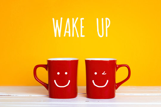 Two red coffee mugs with a smiling faces on a yellow background with the phrase Wake up.