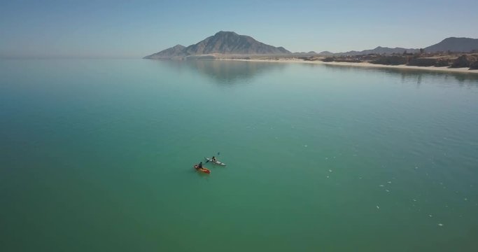 Aerial view of two kayakers paddling on a vast sea with a rocky mountain in the distance.
