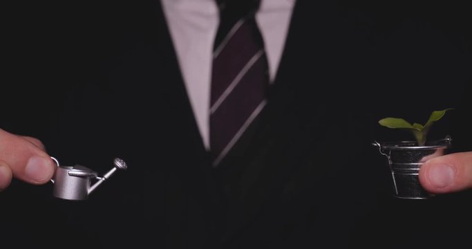 A businessman in a suit holds a sprout in a mini bucket and a watering can, a black background, a career growth, a new life.
