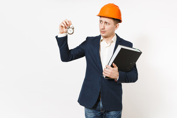 Young concerned businessman in dark suit, protective hardhat holding black folder for papers document and alarm clock isolated on white background. Time is running out. Male worker for advertisement.
