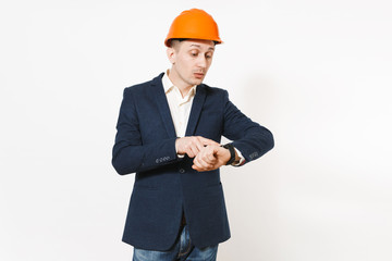 Young concentrated businessman in dark suit, protective construction orange helmet pointing index finger on clock on hand isolated on white background. Time is running out. Male worker, advertisement.