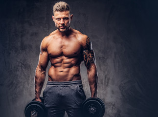 Fototapeta na wymiar A handsome shirtless tattooed bodybuilder with stylish haircut and beard, wearing sports shorts, posing in a studio. Isolated on a dark background