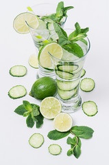 Fresh detox spring cocktail with mint, lime, ice, cucumber, straw on soft white wooden board as fun summer holiday background, top view, closeup.