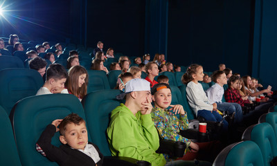 Sideview of boys watching movie in the cinema. There are other people wearing colorful clothes on background. They laughing, smiling, eating popcorn and drinking cola.