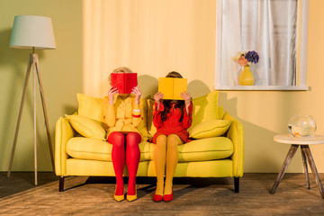 obscured view of women in retro clothing with books sitting on yellow sofa at bright apartment,...