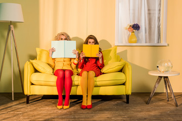 obscured view of women in retro clothing with books sitting on yellow sofa at bright apartment, doll house concept