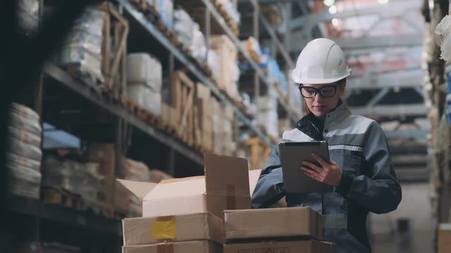 A woman in a warehouse in a white helmet and gray uniform opens a cardboard box, looks at the goods and makes an inventory. Merchandiser recounts the item in the box and makes notes on the tablet.