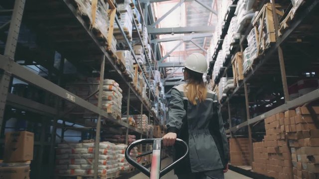 Following shot of female industrial worker in hard hat walking through along shelves with trolley for the warehouse. Female laborer in uniform passing by, pulling trolley stuffed with piled boxes
