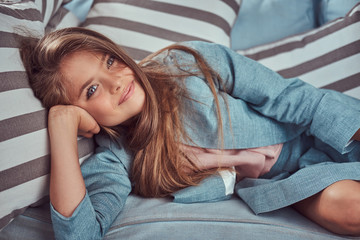 Portrait of a cute little girl with long brown hair, piercing glance and charming smile, looking at a camera, lying on a sofa at home alone