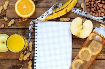 Healthy lifestyle; fitness bottle; fruits (oranges; apples and bananas); hazelnuts and peanuts; orange juice, a pen and a notebook and measuring tape. Top view; the concept of free space for text
