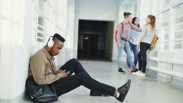 Young handsome african american student sitting on floor in white corridor with headphones on head listening to music holding smartphone texting someone. His classmates are behind him