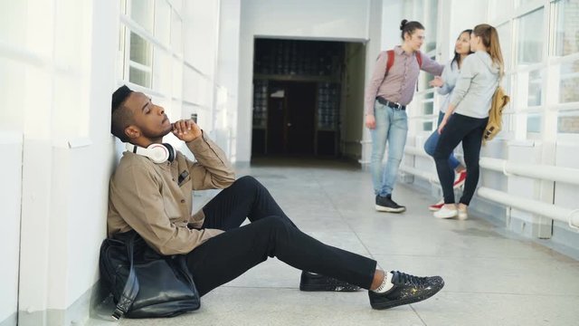 Upset serious african-american male student sitting on floor in university corridor looking in different sides while his groupmates are standing communicating