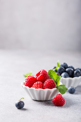 Fresh ripe raspberries and bluebetties in white bowls on light gray concrete background. Healthy food concept with copy space.