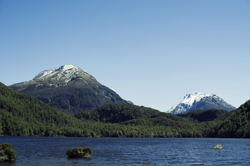 Paisaje de picos de monta&ntilde;as nevados y verdes con cielo azul frente a un lago en Nueva Zelanda