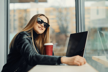 Young girl sit in coffee place in front of the window look at her laptop and drink tea from red cup