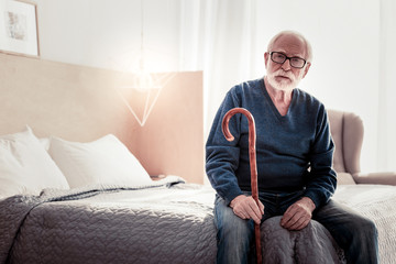 Old age. Serious nice elderly man sitting on the bed an holding his walking stick while resting at home