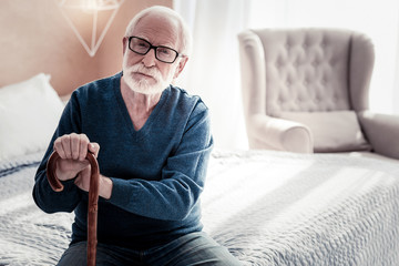 My home. Nice pleasant aged man sitting on the bed and holding his walking stick while resting in the bedroom