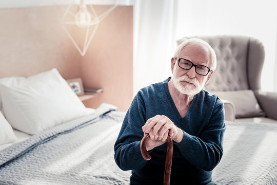 At Home. Pleasant Nice Senior Man Sitting In The Bedroom And Holding His Walking Stick While Resting At Home