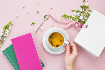 Flatlay hands of a young woman, notebook, cup of tea, white gift bag and spring flowers on a pink background. Beautiful breakfast. Workspace pastel colors. Pink floral background. 