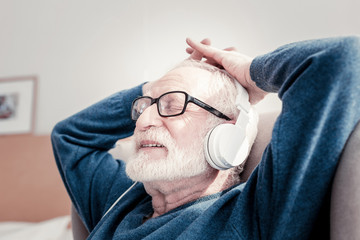 Pleasant pastime. Delighted nice elderly man sitting in the armchair and listening to music while...