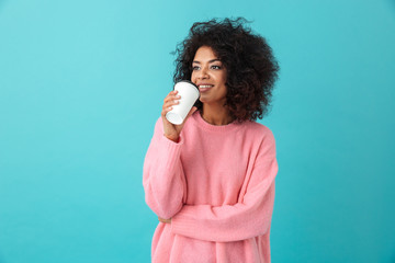 Portrait of american woman 20s with afro hairdo looking aside while drinking takeaway coffee in paper cup, isolated over blue background