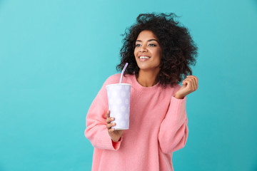 Portrait of adorable woman 20s with afro hairdo looking aside while holding soda beverage in paper cup, isolated over blue background