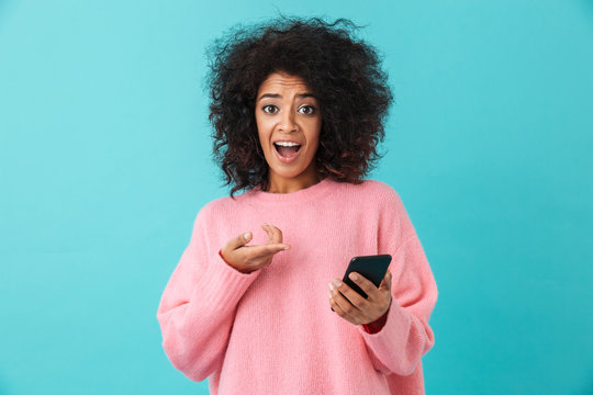 Image of american curly woman in pink shirt reacting emotionally and pointing finger on smartphone, isolated over blue background