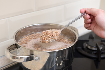 The girl's hand holds cooked buckwheat in a spoon over a saucepan in the kitchen