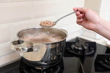 The girl's hand holds cooked buckwheat in a spoon over a saucepan in the kitchen