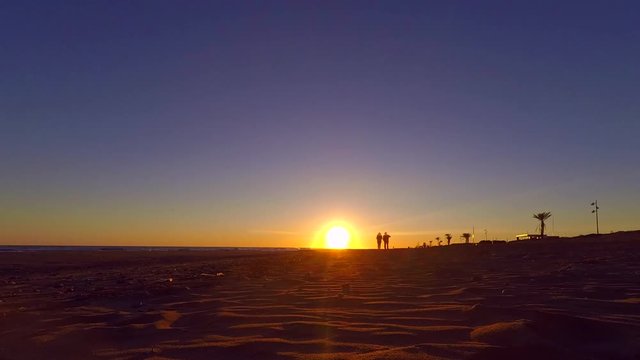 This Is Timelapse Footage Of An Adult Couple Walking On The Beach, They Are Holding Hands And Having A Fun Time Together. Some Planes Can Be Seen Creating Shooting Stars And Leaving Trails